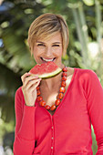 A mature woman with short blonde hair outside wearing a red shirt and holding a slice of watermelon