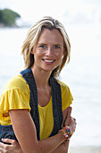 A young blonde woman on a beach wearing a yellow top and a denim waistcoat