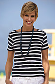 A young blonde woman with short hair on a beach wearing a black-and-white striped top and white summer trousers