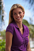 A young blonde woman under palm trees wearing a purple top