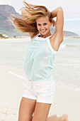 A young blonde woman on a beach wearing a light-blue top and white shorts