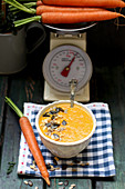 Carrot cream soup in a small bowl on a checked towel, with carrots on a scale in the background