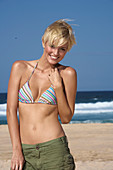 A blonde woman with short hair on a beach wearing a bikini top and shorts