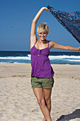 A blonde woman with short hair on a beach wearing a purple top and olive-green shorts