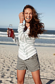A brunette woman on a sandy beach with a drink wearing a striped top and shorts