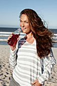 A brunette woman on a sandy beach with a drink wearing a striped top and shorts