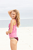 A young blonde woman on a beach wearing a pink top and a bikini