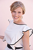 A blonde woman standing against a pink background wearing a light blouse with a black trim