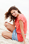 A brunette woman on a beach wearing a salmon-coloured top and a denim skirt