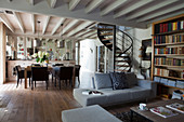 Grey sofa set and bookcase in front of spiral staircase in open-plan interior with dining area in background