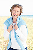 Reife Frau mit blonder Kurzhaarfrisur in Bluejeans und weißem Shirt mit blauem Pullover über der Schulter in der Natur