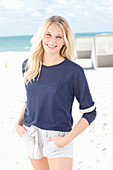 A young blonde woman on a beach wearing a long-sleeved blue shirt and shorts