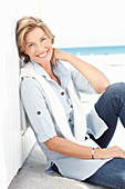 A mature woman with short blonde hair on a beach wearing a light-blue striped short, jumper over her shoulders and blue jeans