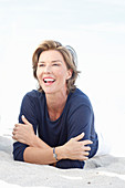 A mature woman with short blonde hair on a beach wearing a blue shirt and white trousers