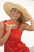 A young blonde woman wearing a red summer dress and a summer hat holding a slice of watermelon
