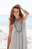 A young brunette woman on a beach wearing a beige summer dress and a necklace