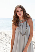 A young brunette woman on a beach wearing a beige summer dress and a necklace