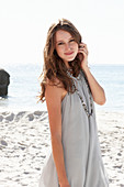 A young brunette woman on a beach wearing a beige summer dress and a necklace