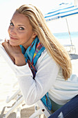A blonde woman on a beach wearing a colourful scarf and a white jacket