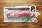 A raw pork fillet with various ingredients and a knife on a wooden background