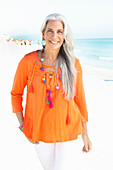 A mature woman with white hair on a beach wearing an orange tunic and white summer trousers