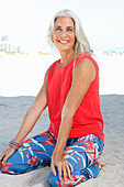 A mature woman with white hair on a beach wearing a red top and a pair of floral-patterned summer trousers