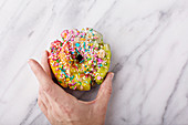 Colorful and festive unicorn donut with sprinkles on marble surface with a woman s hand holding it, unicorn food trend
