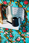 Vase, small watering can and tray on white wall shelf in front of floral wallpaper