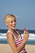 A blonde woman with short hair by the sea wearing a red-and-white striped top and holding a slice of watermelon