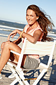 A brunette woman wearing an apricot blouse and shorts sitting on a chair by the sea