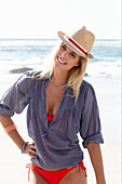 Blonde Frau mit Hut in rotem Bikini und blauer Bluse am Strand