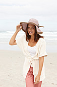 A brunette woman by the sea wearing a short-sleeved cardigan and a hat