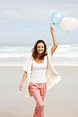 A brunette woman by the sea wearing a short-sleeved cardigan and holding balloons