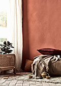 Kissen, Decke und Teegeschirr auf Teppichvorleger vor ziegelroter Wand, Zimmerpflanze vor Fenster