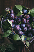 Damsons with leaves in a container on a dark wooden surface