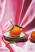 Mandarine mit Blatt auf Tellerstapel vor pinkem Stoffhintergrund
