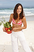 A young brunette woman on a beach wearing a bikini top and white trousers and holding a bowl of vegetables