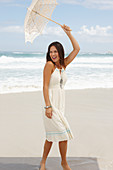 A brunette woman by the sea wearing a white summer dress and holding a parasol