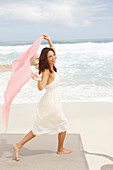 A brunette woman by the sea wearing a white summer dress and holding a pink shawl