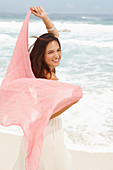 A brunette woman by the sea wearing a white summer dress and holding a pink shawl