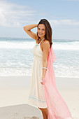 A brunette woman by the sea wearing a white summer dress and holding a pink shawl