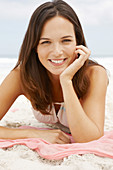 A brunette woman lying on a pink shawl by the sea wearing a white summer dress