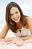 A brunette woman lying on a pink shawl by the sea wearing a white summer dress