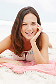 A brunette woman lying on a pink shawl by the sea wearing a white summer dress