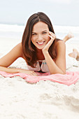 A brunette woman lying on a pink shawl by the sea wearing a white summer dress