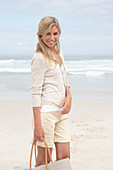 A blond woman on a beach wearing a light cardigan and shorts and holding a bag