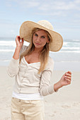 A blond woman on a beach wearing a light cardigan, shorts and a summer hat