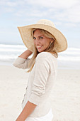 A blond woman on a beach wearing a light cardigan and a summer hat