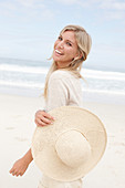 A blond woman on a beach wearing a light cardigan and a summer hat