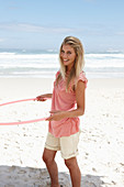 A young woman on a beach with a hula-hoop wearing a pink top and shorts